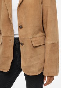 Tan suede blazer with a relaxed fit, large pockets, and two button closures. Matte texture with visible stitching details. Designed for layering.