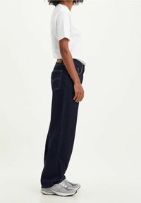Pantalones vaqueros de mezclilla azul oscuro con corte recto, que cuentan con dos bolsillos traseros, costuras cosidas y una cintura alta, combinados con una camiseta blanca y zapatillas grises.