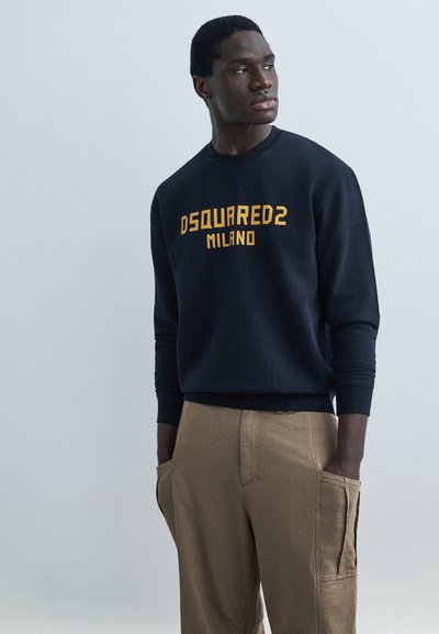 Mann ikledd en svart genser med teksten «DSQUARED2 MILANO» i gult, brune bukser med store sidelommer, står med hendene i lommene og ser mot høyre.