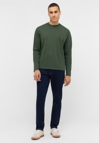 Koszula z długim rękawem w oliwkowym zielonym kolorze z okrągłym dekoltem i subtelnym logo, w połączeniu z ciemnoniebieskimi dżinsami i beżowymi sneakersami.