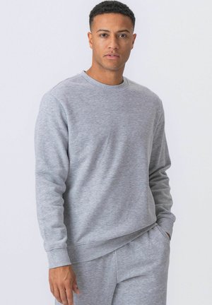 Mężczyzna ubrany w jasnoszarą bluzę z okrągłym dekoltem i pasujące spodnie dresowe, stojący z jedną ręką w kieszeni na jednolitym tle.