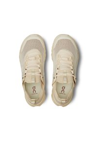 Chaussures de sport beige clair avec une tige en mesh, bout rond, lacets souples et semelles en caoutchouc proéminentes. Logo de la marque visible sur la languette.