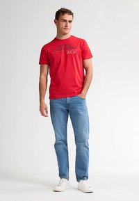 Tricou roșu din bumbac cu un logo negru imprimat „PETROL INDUSTRIES & CO” pe partea din față, asortat cu blugi din denim albastru deschis și adidași albi.