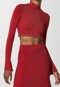 Červený zkrácený svetr s dlouhými rukávy, pružným žebrováním, vysokým výstřihem a stříbrnými kovovými doplňky podél spodního lemu. V kombinaci s odpovídající sukní.