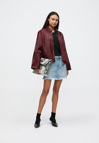 Veste en daim bourgogne avec un col montant et des détails en anneau, associée à un T-shirt noir et à une jupe en denim clair. Les accessoires incluent une pochette en peau de serpent.