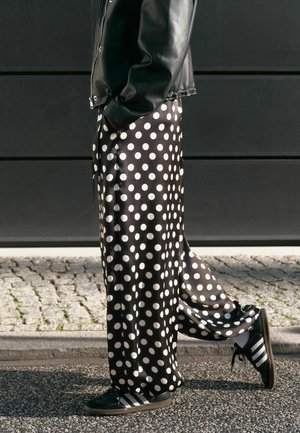 Isik, kes kannab musta nahktagi, musti laia säärtega pükse valgete täppidega ja musti tossusid valgete triipudega, seisab tänaval.