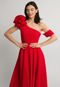 Czerwona sukienka na jedno ramię z dużym falbaną na jednym rękawie, gładki materiał i rozkloszowany dół, który opada na ziemię.