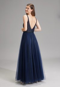 Donna che sorride sopra la spalla, indossando un abito senza maniche blu navy con profondo scollo a V e gonna in tulle ampia, in piedi su uno sfondo grigio chiaro.
