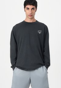 Zwarte lange mouwen shirt van katoen, met een ronde hals en wit logo op de borst. Draagt bij lichte grijze shorts.