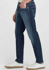 Jeans de mezclilla azul oscuro con corte recto, cinco bolsillos y costuras sutiles. Combinados con zapatillas blancas con suela marrón.