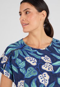 Top a maniche corte con sfondo blu scuro e stampa di foglie tropicali in verde e crema. Presenta un collo rotondo e cuciture decorative.