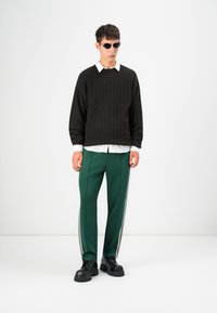 Pullover ribbed maro �închis peste o cămașă albă cu guler, asortat cu pantaloni de trening verzi cu dungi laterale albe și încălțăminte chunky neagră.