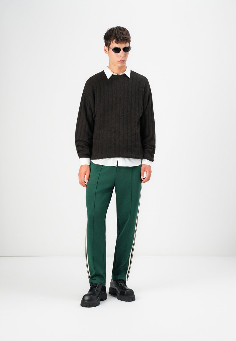 Pull en côtes brun foncé sur une chemise à col blanc, associé à un pantalon de survêtement vert avec des rayures blanches sur les côtés et des chaussures noires épaisses.