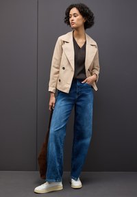 Kobieta z kręconymi włosami nosi beżową kurtkę, czarny sweter w serek, szerokie niebieskie dżinsy, białe trampki i trzyma brązową torbę na szarym tle.