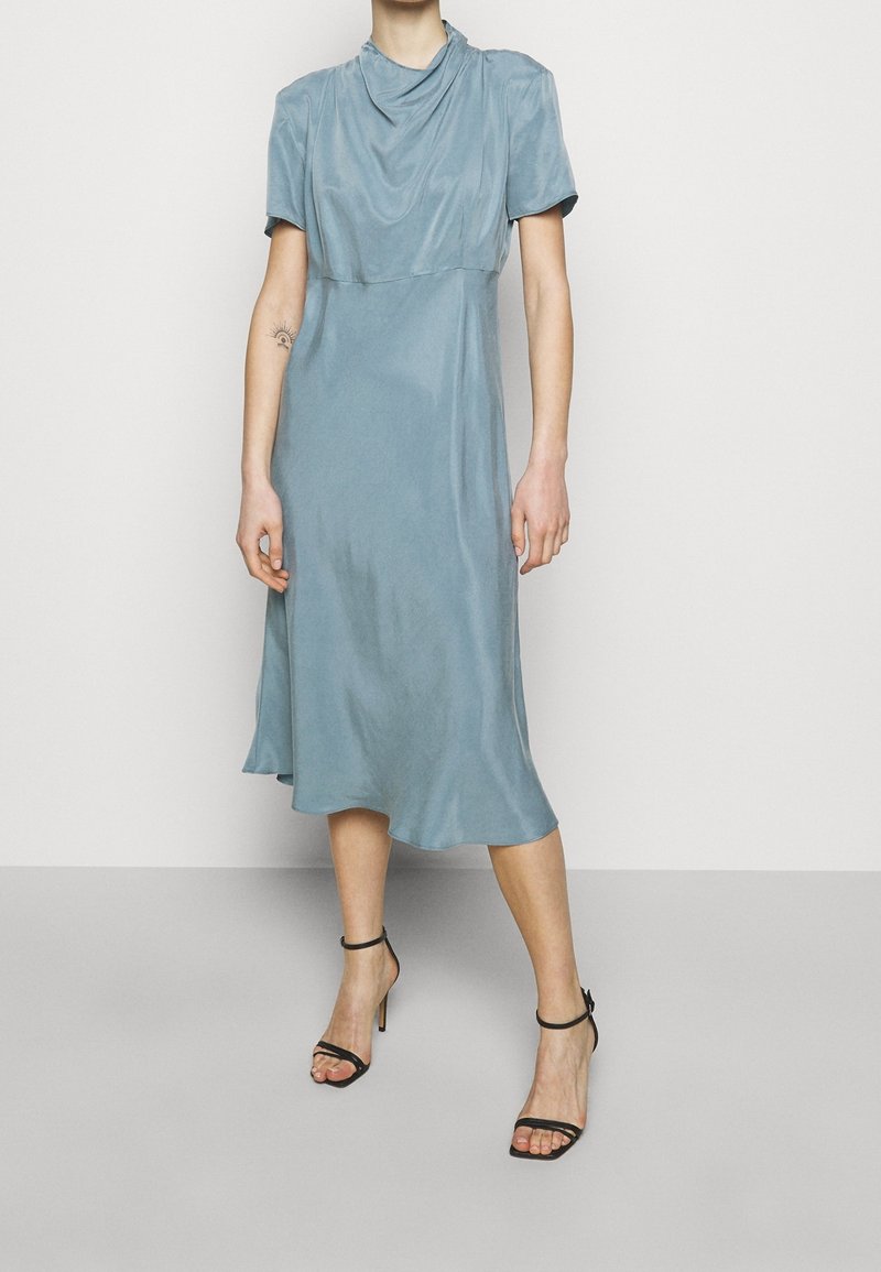 Kvinna som bär en ljusblå midiklänning med korta ärmar och svarta remsandaler med hög klack, stående mot en vanlig bakgrund.