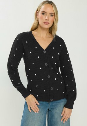 Kvinde iført sort knap-op cardigan med små hvide prikker og blå jeans, poserer med den ene hånd på hoften mod en ensfarvet baggrund.