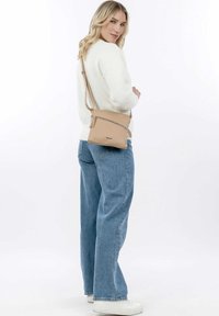 Béžová kožená taška na crossbody s přední kapsou na zip, obdélníkový tvar a nastavitelný popruh, nošena s bílým svetrem a modrými džíny.