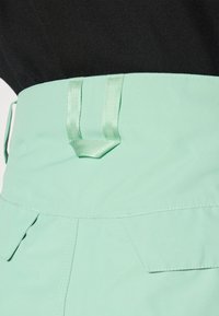 I pantaloni verde menta presentano una cintura ampia con due asole per nastri, tasche laterali e dettagli di cucitura visibili; il tessuto è liscio e opaco.