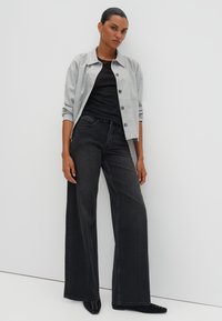 Veste grise à boutons sur un haut noir, associée à un jean noir à jambes larges. Cette tenue présente une coupe décontractée et des textures de tissu lisses.