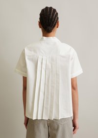 Hvit kortermet skjorte med plissert bak, som har krage og avslappet passform. Stoffet virker glatt og lett.