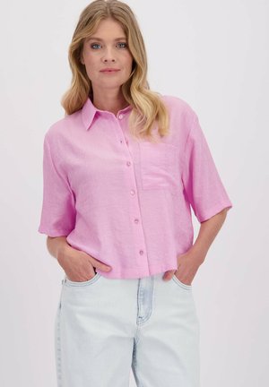 Lichtroze linnen overhemd met korte mouwen, een kraag, knoopsluiting aan de voorkant en een enkele borstzak, gedragen met vervaagde lichtblauwe jeans.