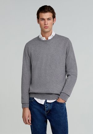 Sudadera gris de cuello redondo hecha de tela suave, con puños y dobladillo acanalados, combinada con una camisa blanca de cuello y jeans azules.