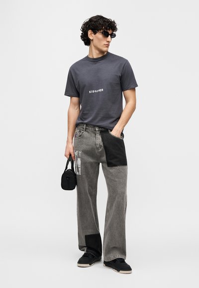 Γκρι βαμβακερό μπλουζάκι με το λογότυπο "KID SUPER", συνδυασμένο με τζιν παντελόνι patchwork γκρι με μαύρες λεπτομέρειες και γραφικά, φορεμένο με μαύρα παπούτσια.