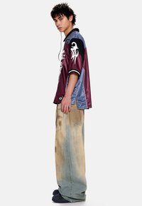 Chemise oversize à manches courtes bordeaux et bleue avec des motifs graphiques, associée à un pantalon bleu délavé ample et des chaussures noires.