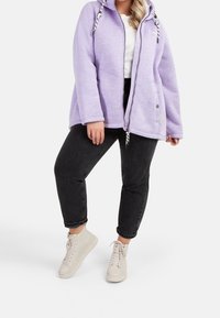 Fioletowa kurtka z kapturem i zamkiem wykonana z miękkiego materiału, z czarno-białymi sznurkami; zestawiona z czarnymi jeansami z podwiniętymi nogawkami i jasnymi butami sportowymi.