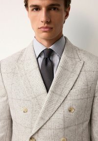 Lys beige dobbeltbrystet blazer med struktureret stof, prydet med et subtilt mønster. Underneath en lyseblå stribet skjorte og et mønstrede slips.
