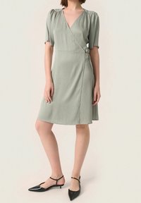 Světlounce zelené šaty ve stylu zavinování s krátkými nabíranými rukávy, texturovaným materiálem a zavazováním na boku. Kombinované s černými botami se špičatou špičkou.