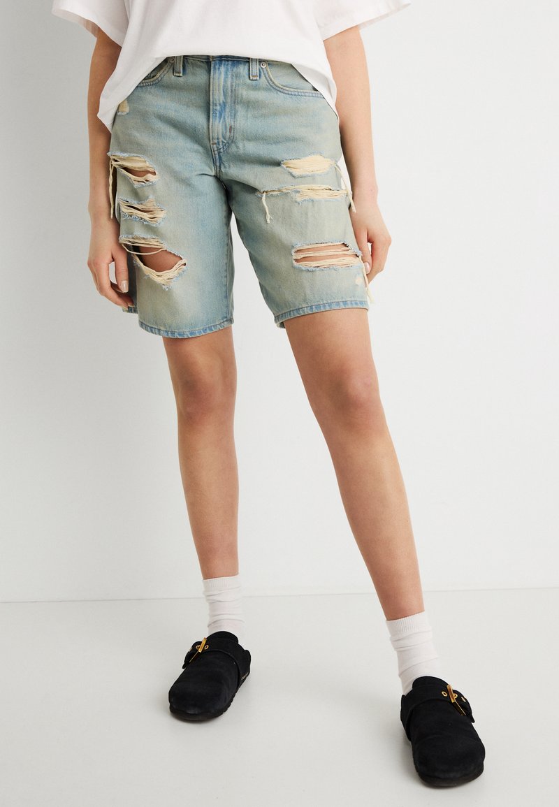 Άτομο που φοράει ανοιχτόχρωμα σκισμένα τζιν σορτς, λευκές κάλτσες, μαύρα παπούτσια χωρίς κορδόνια και άνετο λευκό πουκάμισο, στέκεται μπροστά σε λευκό φόντο.