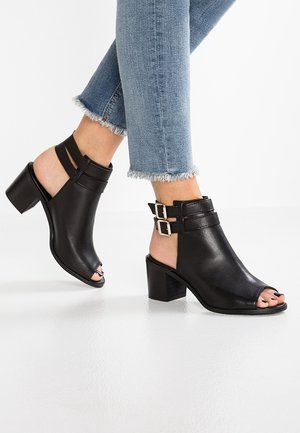 Kvinde iført sorte læder åbne-til-tå hælsandaler med dobbelte ankelstropper og lyseblå jeans med frynset kant.