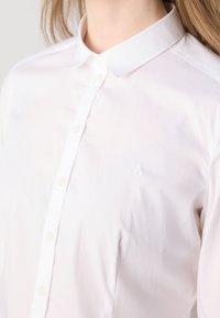 Femme portant une chemise blanche cintrée à boutons avec un col pointu et un logo brodé discret sur la poitrine.