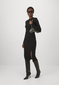 Melna garā piedurknē kleita ar sānu izgriezumu, apvienota ar melniem zābakiem līdz ceļiem un zaļu rokassomu. Iezīmēta ar pieguļošu siluetu un gludu audumu.