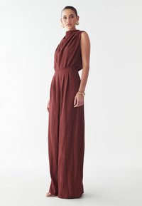 Punane, ilma varrukat jumpsuit kõrge kaelusega, laiade jalgade ja tekstuurse kangaga. Model kannab kuldseid kõrvarõngaid ja käevõru, seistes neutraalses taustas.