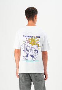 Weißes Baumwoll-T-Shirt mit einem Grafikmotiv einer Cartoon-Takeout-Box, gefüllt mit Nudeln und Essstäbchen. Der Text lautet "Chinatown" in Lila und Türkis.
