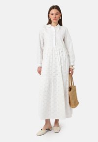 White long-sleeve dress with a button-up front, featuring textured eyelet detailing, a tiered skirt, and casual slip-on shoes.