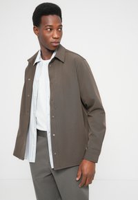 Καφέ σακάκι με κουμπιά και γιακά, φορεμένο πάνω από πουκάμισο με ανοιχτό μπλε ρίγες, συνδυασμένο με γκρι παντελόνι. Λείο ύφασμα, casual σχεδιασμός.