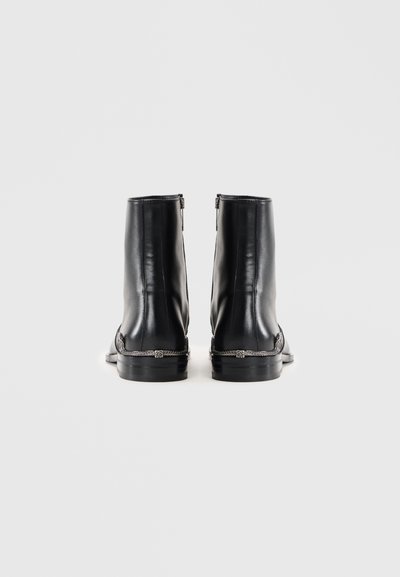 Ett par svarta läderankelboots med låga klackar och silvriga kedjedetaljer, visade från baksidan mot en enfärgad vit bakgrund.