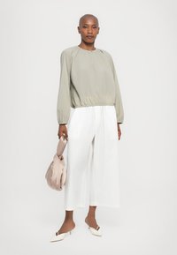 Blusa lunga verde chiaro con orlo arricciato; abbinata a pantaloni bianchi larghi. La modella tiene una borsa morbida color beige. Le scarpe sono mule bianche.
