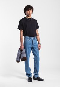 Černé tričko s krátkým rukávem, světle modré rovné džíny, držící vzorovaný svetr, s černými botami, stojící na bílém pozadí.