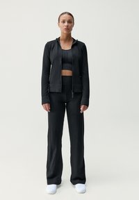 Svart atletisk set som består av en åtsittande crop top med perforerat mönster och en jacka med hel dragkedja. Lösa svarta byxor, kombinerade med vita skor.