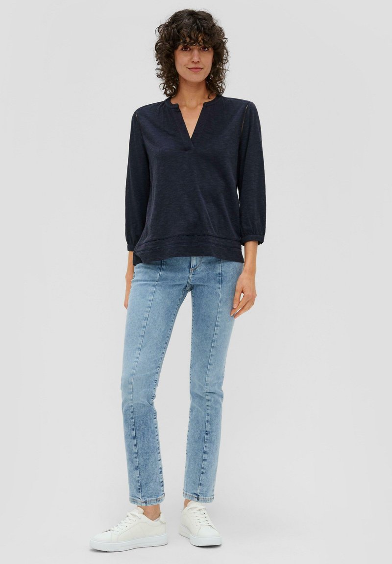 Marineblauer Pullover mit V-Ausschnitt und Dreiviertelärmeln, kombiniert mit hellblauen Skinny-Jeans und weißen Sneakers. Weiche Textur, lässiger Schnitt.