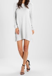 Abito grigio a maniche lunghe realizzato in tessuto morbido, con una vestibilità rilassata, orlo arrotondato e dettagli laterali, abbinato a tacchi neri con cinturini.