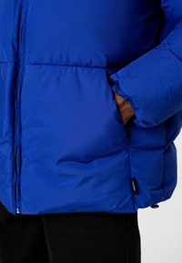 Blauwe pufferjas met een glanzende afwerking, voorzien van een rits, elastische manchetten en een zijzak, tegen een neutrale achtergrond gepresenteerd.