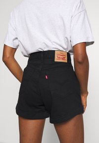 Shorts di denim nero a vita alta con orli risvoltati, caratterizzati da una toppa in pelle marrone sul retro e da una piccola etichetta rossa sulla tasca.