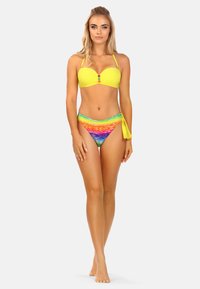 Kobieta idąca boso, ubrana w żółty stanik bikini ozdobiony koralikami oraz bikini w tęczowe paski, z żółtym wiązaniem po lewej stronie.