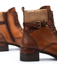 Botas de tobillo de cuero en marrón con un acabado texturizado. Presentan un puño de tela acanalada, cremallera lateral y un acento decorativo de hebilla.