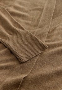 Maglione marrone con una texture liscia, caratterizzato da un polsino a costine. Il tessuto è morbido e sembra leggero, con un design aperto.